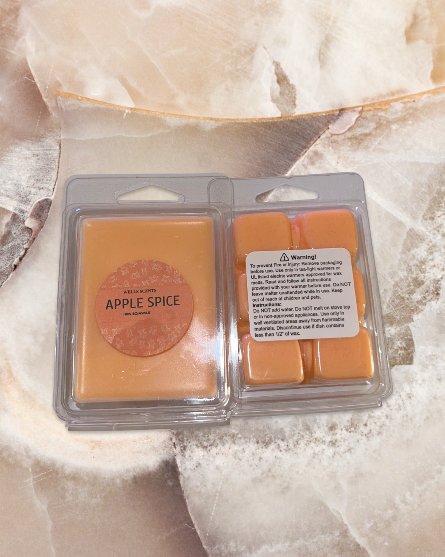 Apple Spice scented wax melts in amber color, packaged in clear plastic clamshell containers with two sections, displayed on a cream marble surface. Product features a circular label with warm autumn-inspired design.